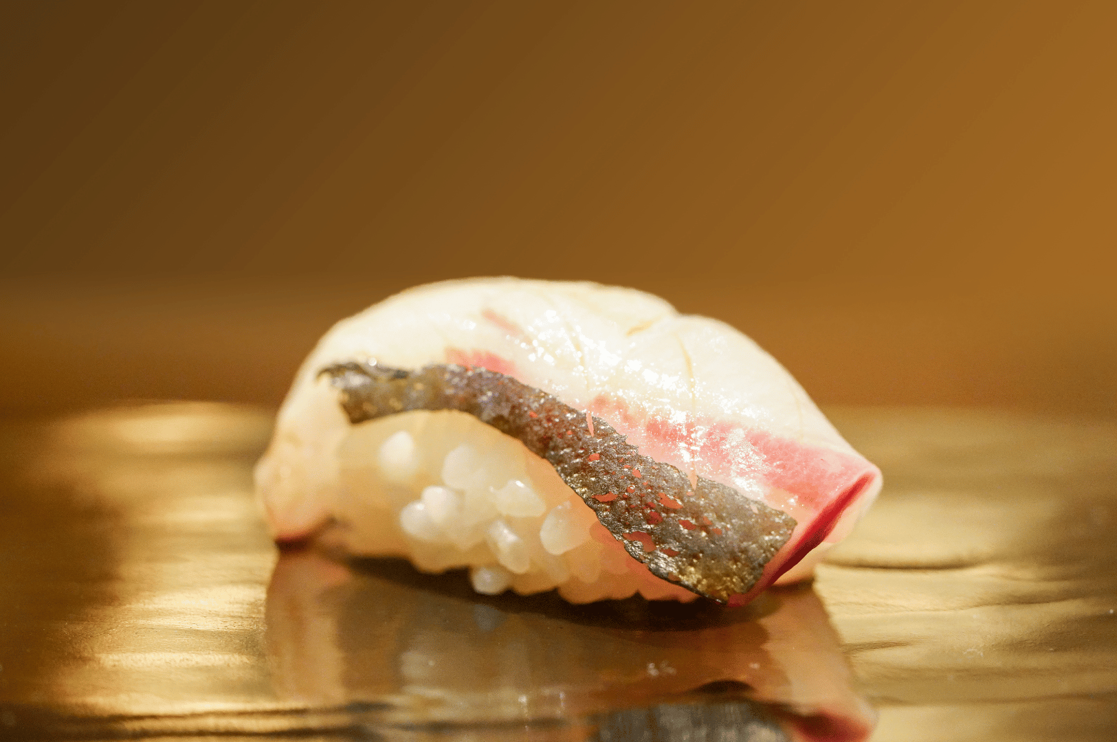 A single piece of glistening nigiri rests on a reflective surface, its silver-skinned fish catching the warm ambient light. The minimalist presentation invites quiet contemplation, marking the moment when the chef’s craft meets the guest’s anticipation. This pause between preparation and tasting embodies the heart of omakase—trust, timing, and the beauty of restraint.