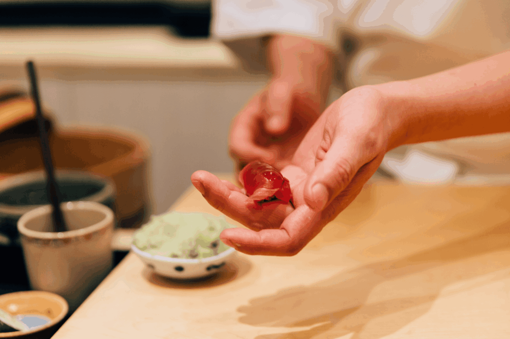 A sushi chef’s hands cradle a piece of tuna nigiri with quiet precision, the gesture intimate and intentional. Surrounding bowls of wasabi and sauces hint at the layered flavors to come, while the warm wood counter evokes the quiet rhythm of omakase service. This moment captures the chef’s silent dialogue with the diner—an offering of trust, craft, and care.