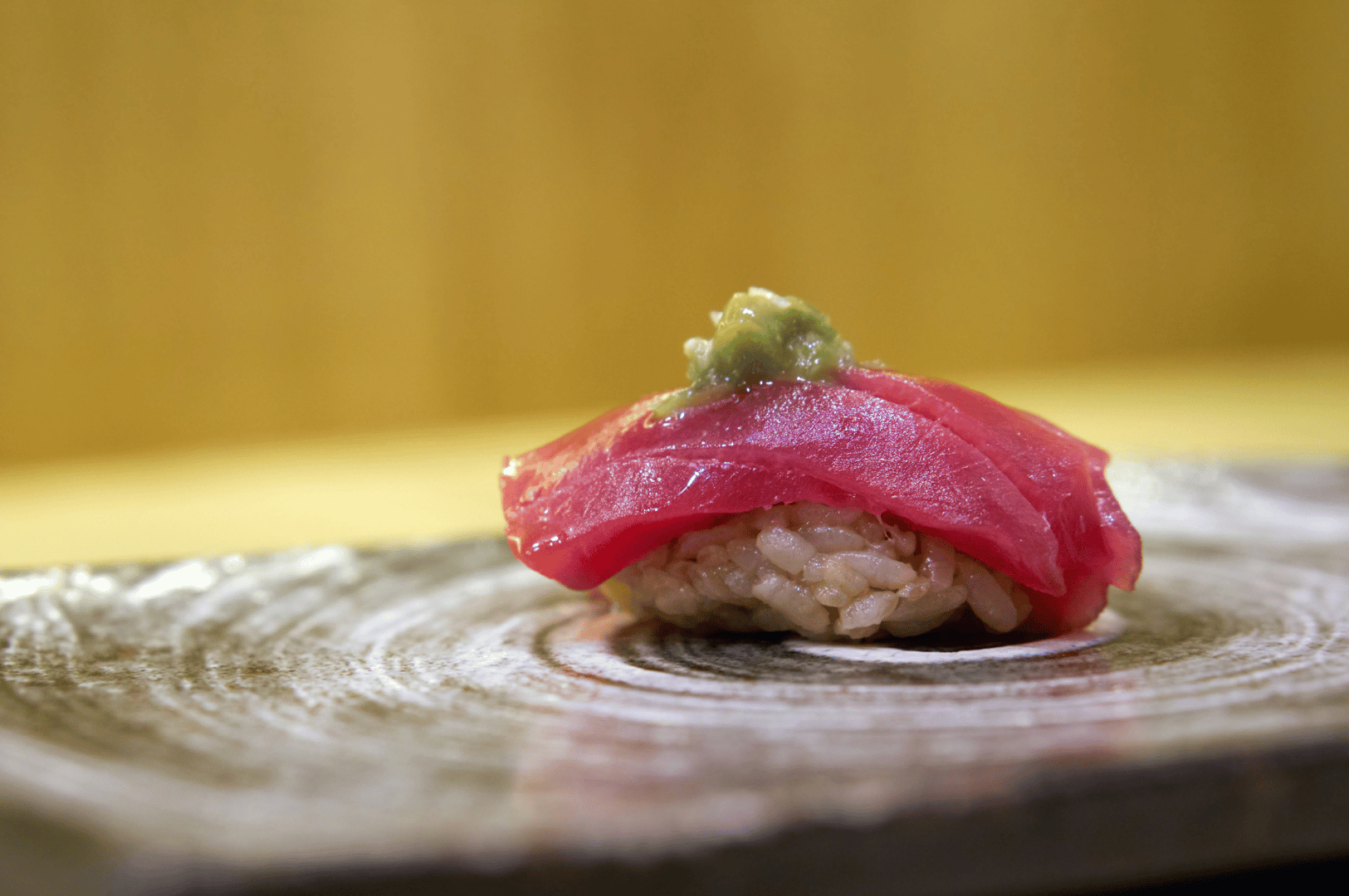 A pristine piece of tuna nigiri at Shinji, placed on a warm-toned wooden surface with a delicate dab of wasabi atop the glistening fish. The softly blurred golden background draws focus to the sushi’s purity and balance—underscoring Shinji’s philosophy of quiet precision and ingredient-led storytelling in contrast to Ginza Sushi Ichi’s theatrical craftsmanship.