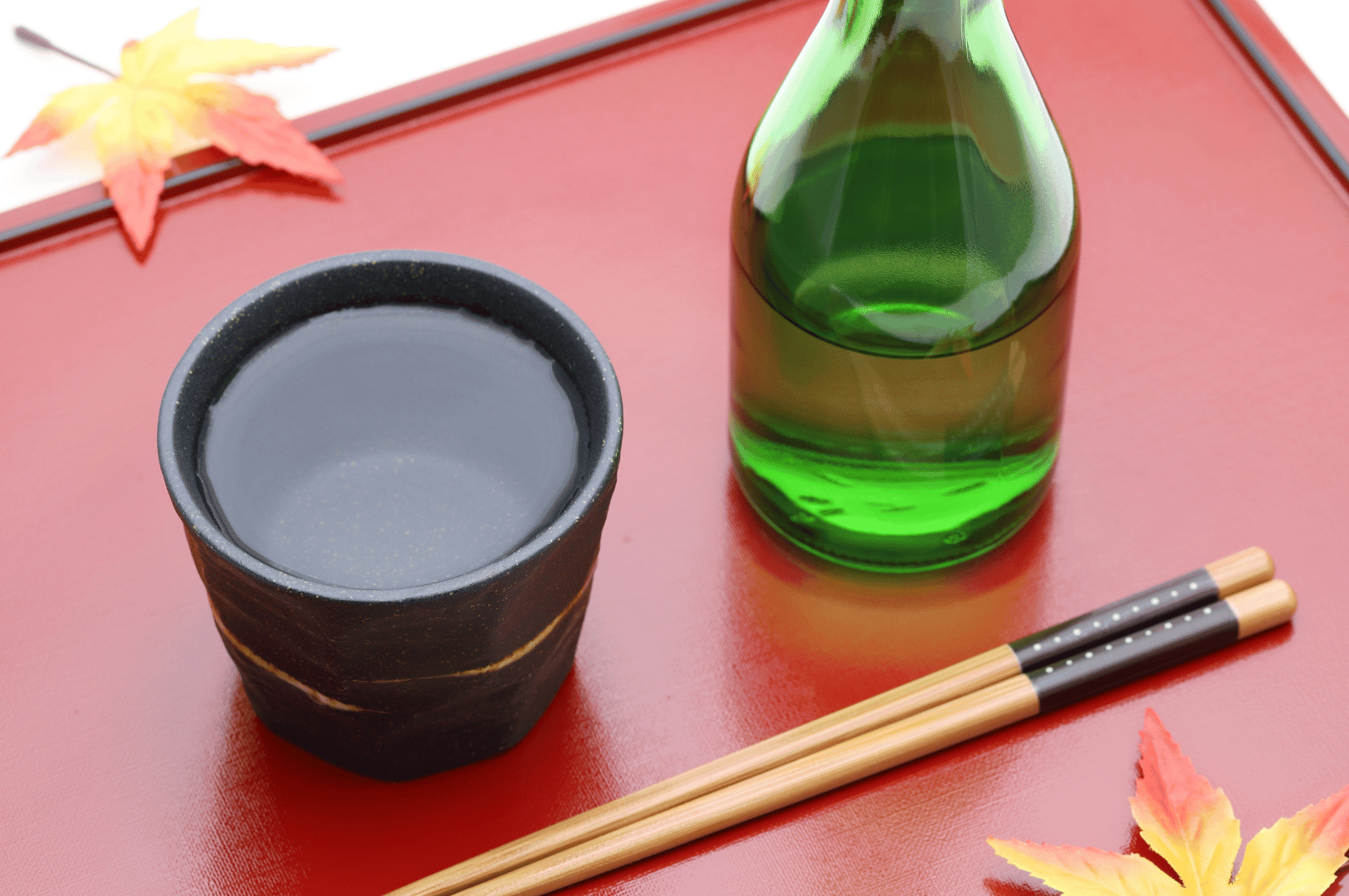 Green sake bottle and black ceramic cup rest on a glossy red lacquer tray, framed by golden-accented chopsticks and vibrant autumn leaves in red, orange, and yellow. The seasonal composition evokes the warmth and reflection of post-celebration rituals, where premium sake and digestif offerings in Singapore elevate Japanese cuisine into an immersive, sensory farewell.