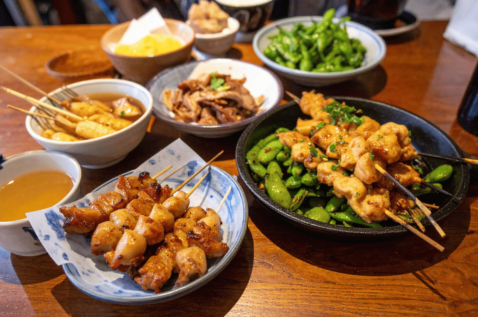 A vibrant spread of Japanese izakaya-style dishes—grilled yakitori with and without green peppers, edamame, simmered meat, oden with daikon and fish cakes, pickled vegetables, and a cup of tea—served on a wooden table, celebrating the communal spirit of Japanese dining while inviting reinterpretation through Singapore’s bold, ingredient-driven fusion.