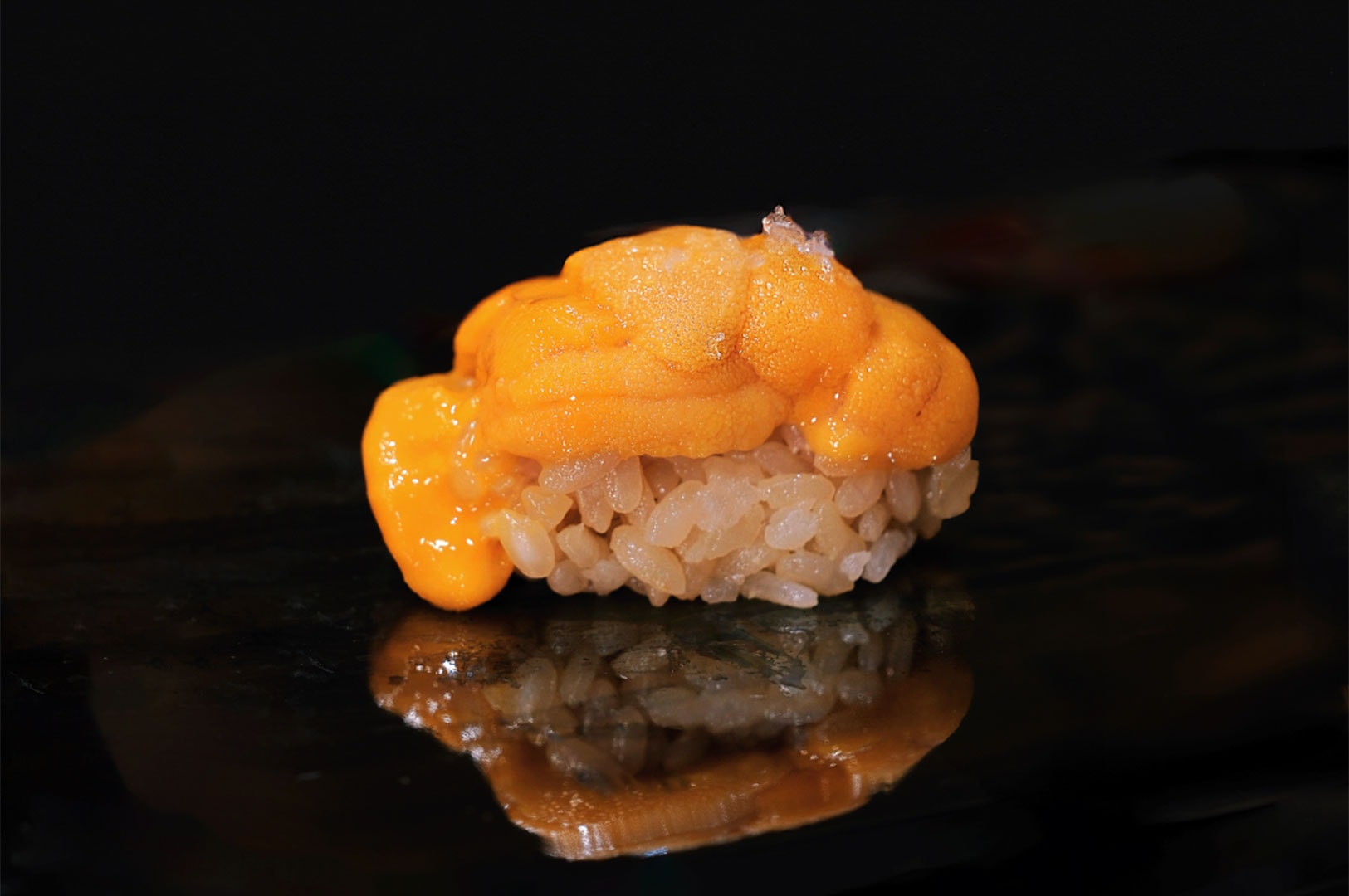 A high-contrast close-up of fresh, bright orange Uni (Sea Urchin) nigiri sushi resting on a dark, reflective surface.