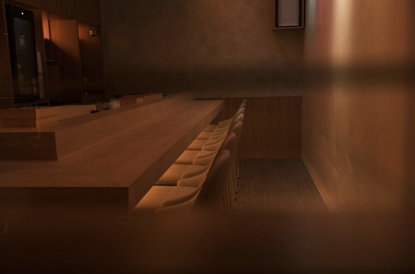 A long wooden bar with several chairs, illuminated softly in a dark setting of a Real Sushi Omakase restaurant.