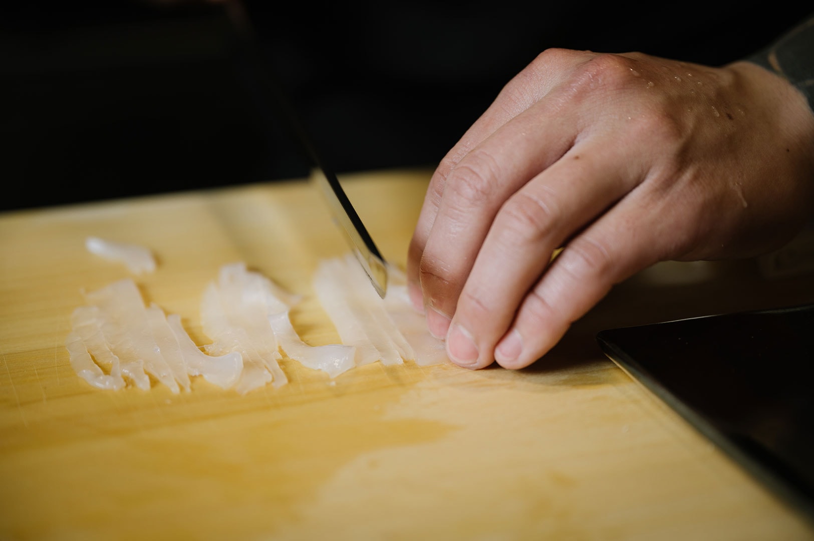 Close-up of a chef's hand using a sharp Japanese knife to precisely slice translucent white fish on a wooden cutting board.