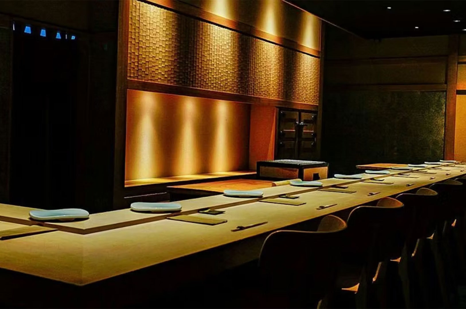 A dimly lit, high-end sushi bar counter featuring warm spotlighting and textured wooden wall panels.
