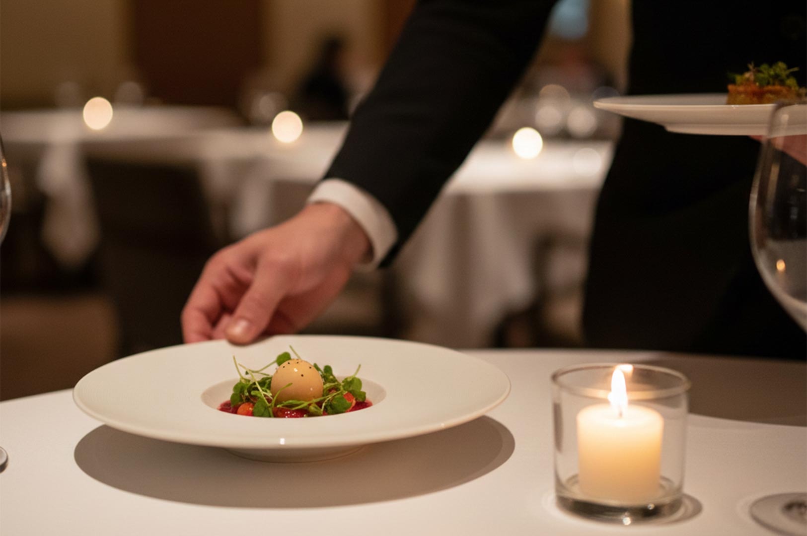 A waiter's hand placing a beautifully plated gourmet appetizer onto a white tablecloth next to a lit candle.