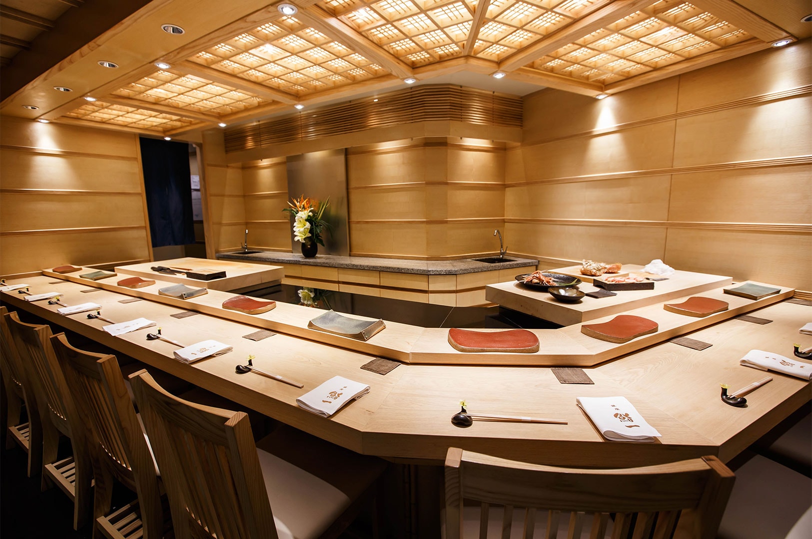 A bright, traditional Japanese sushi restaurant interior featuring light wood paneling, an L-shaped counter, and a coffered ceiling.