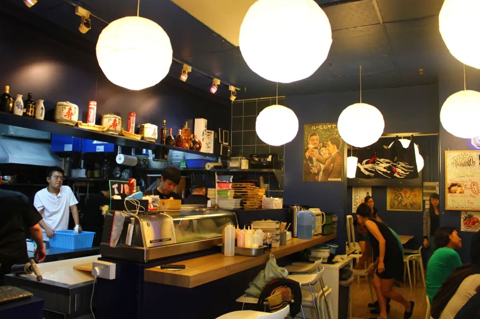 The busy interior of a casual Japanese Izakaya featuring large white paper lanterns hanging from a blue ceiling.