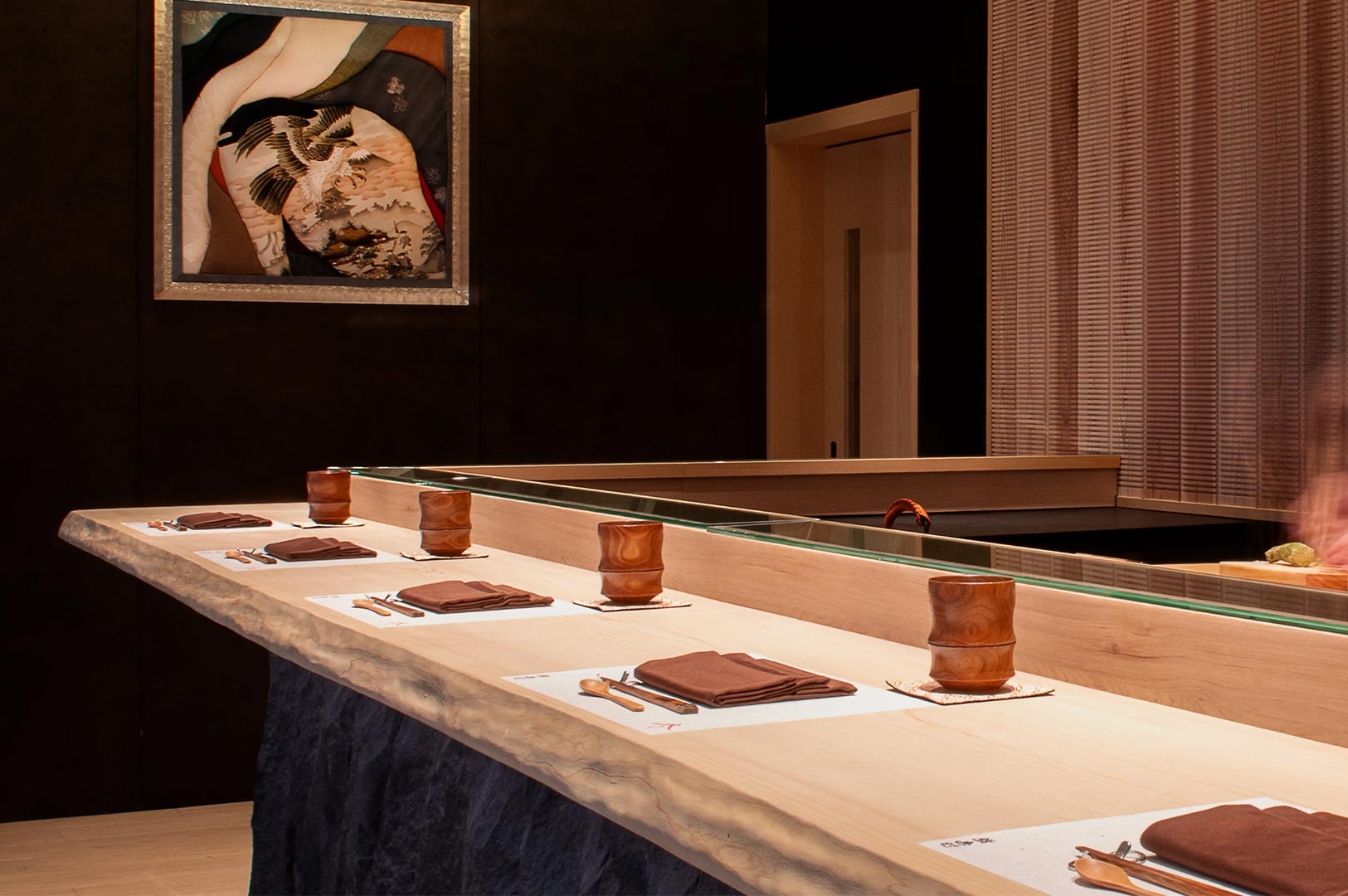 The interior of a high-end sushi restaurant showing a live-edge wooden counter, elegant place settings with wooden cups, and Japanese artwork on a dark wall.