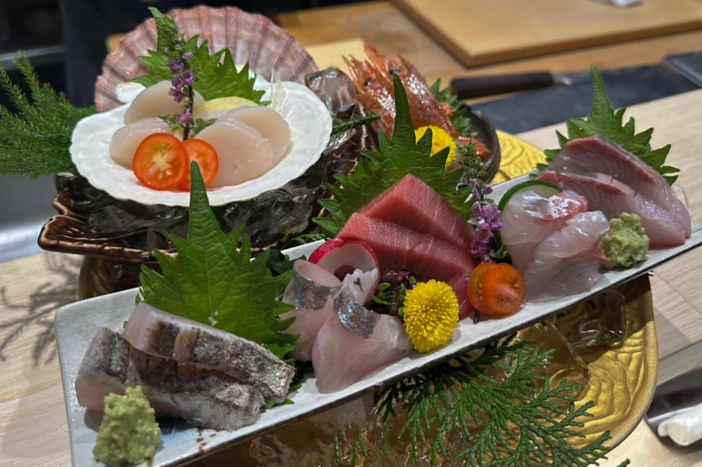 Shot of an elaborate sashimi platter served on a long, rectangular white ceramic plate. The arrangement features various cuts of fresh raw fish, including translucent white fish, deep red tuna, and seared skin-on pieces. In the background, a large scallop shell holds fresh, sliced scallops garnished with halved cherry tomatoes. The platter is decorated with bright green shiso leaves, delicate purple flowers, and a yellow chrysanthemum. The dish is presented on a textured gold decorative plate, set against a light wood sushi counter.