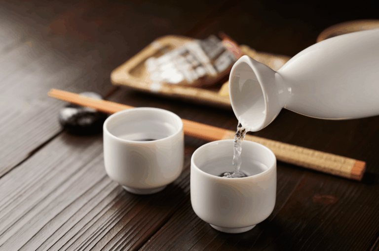 Close-up of a white ceramic tokkuri pouring premium sake into a matching ochoko on a dark wooden table, evoking the quiet elegance of post-celebration rituals. In the background, a bamboo mat, chopsticks, and artisanal packaged bites suggest a curated digestif program. The scene captures the refined intimacy of Japanese dining culture, aligning with Singapore’s elevated sake and digestif offerings that extend the culinary experience beyond the final course.