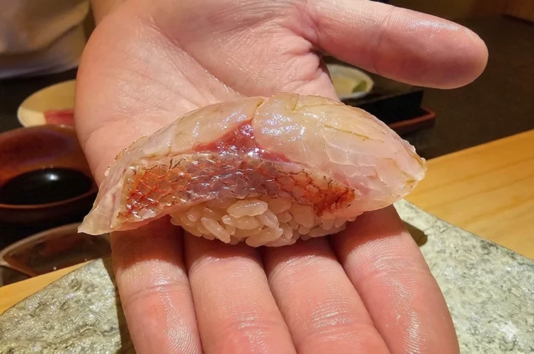 A close-up, eye-level photograph captures a single piece of kinmedai (splendid alfonsino) nigiri resting in the center of an open, upturned palm. The sushi features a thick, translucent slice of pale pink fish with its signature vibrant reddish-orange skin still attached, which has been delicately scored and lightly torched or glazed to a glistening sheen. This succulent cut is draped over a small, compact mound of seasoned white rice, showcasing the contrast between the pearly grains and the textured, colorful fish. In the soft-focus background, a light-colored stone serving board and a warm wooden counter suggest an intimate, high-end omakase setting, with the focused lighting emphasizing the fresh, fatty texture of the premium fish.